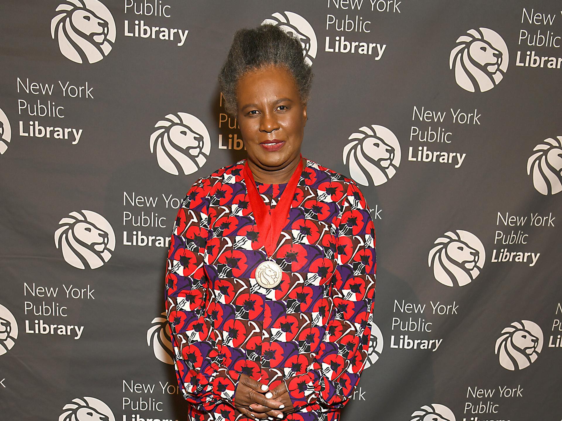 Claudia Rankine lächelt in die Kamera. Sie trägt ein langes Kleid mit rot-blauem Blumenmuster.