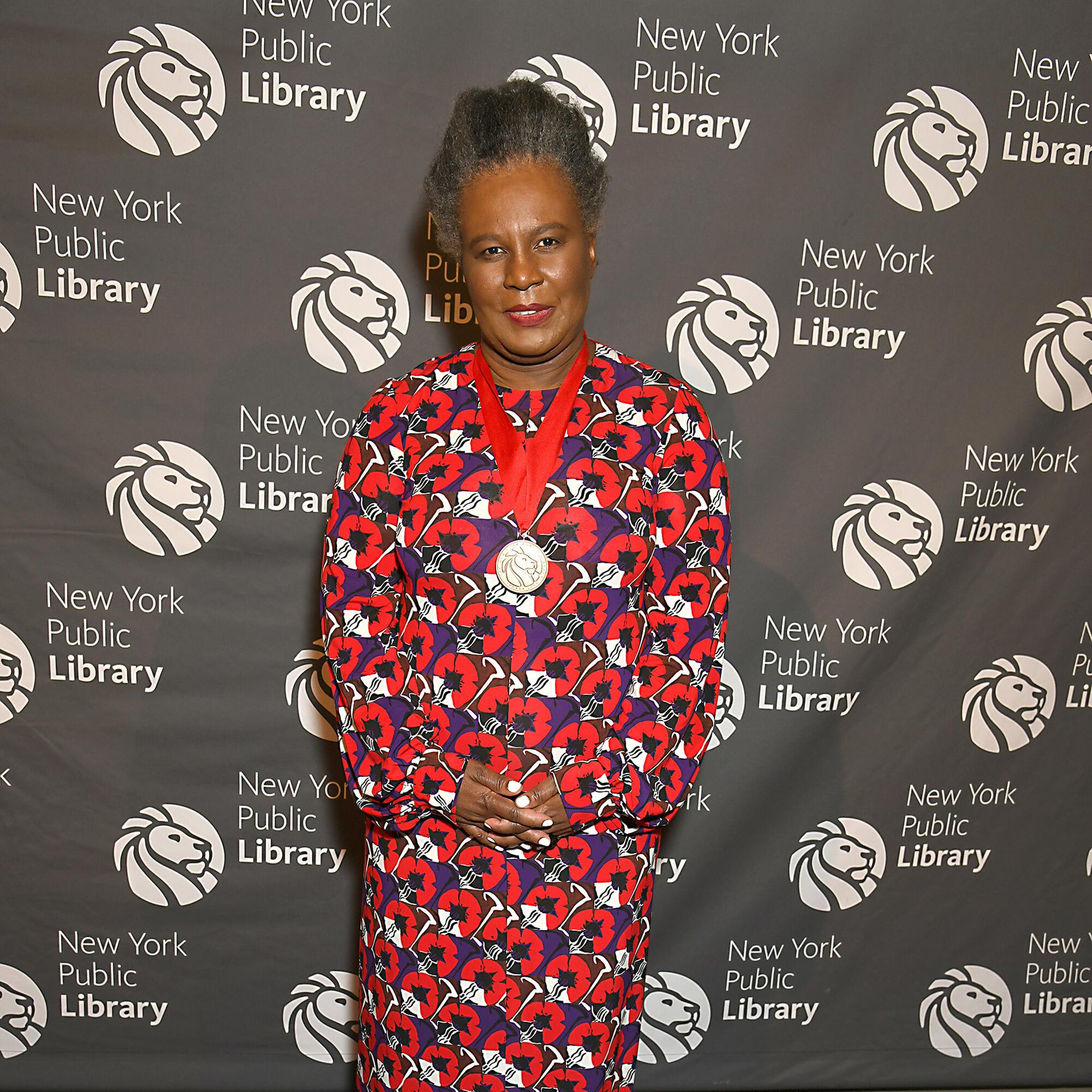 Claudia Rankine lächelt in die Kamera. Sie trägt ein langes Kleid mit rot-blauem Blumenmuster.
