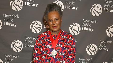 Claudia Rankine lächelt in die Kamera. Sie trägt ein langes Kleid mit rot-blauem Blumenmuster.