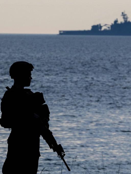 Ein bewaffneter Soldat steht am Strand, in der Ferne ist ein Kriegsschiff sichtbar.