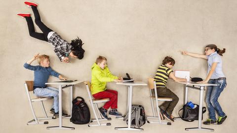 Schulkinder auf Stühlen und an Tischen schweben durch eine Klasse.
