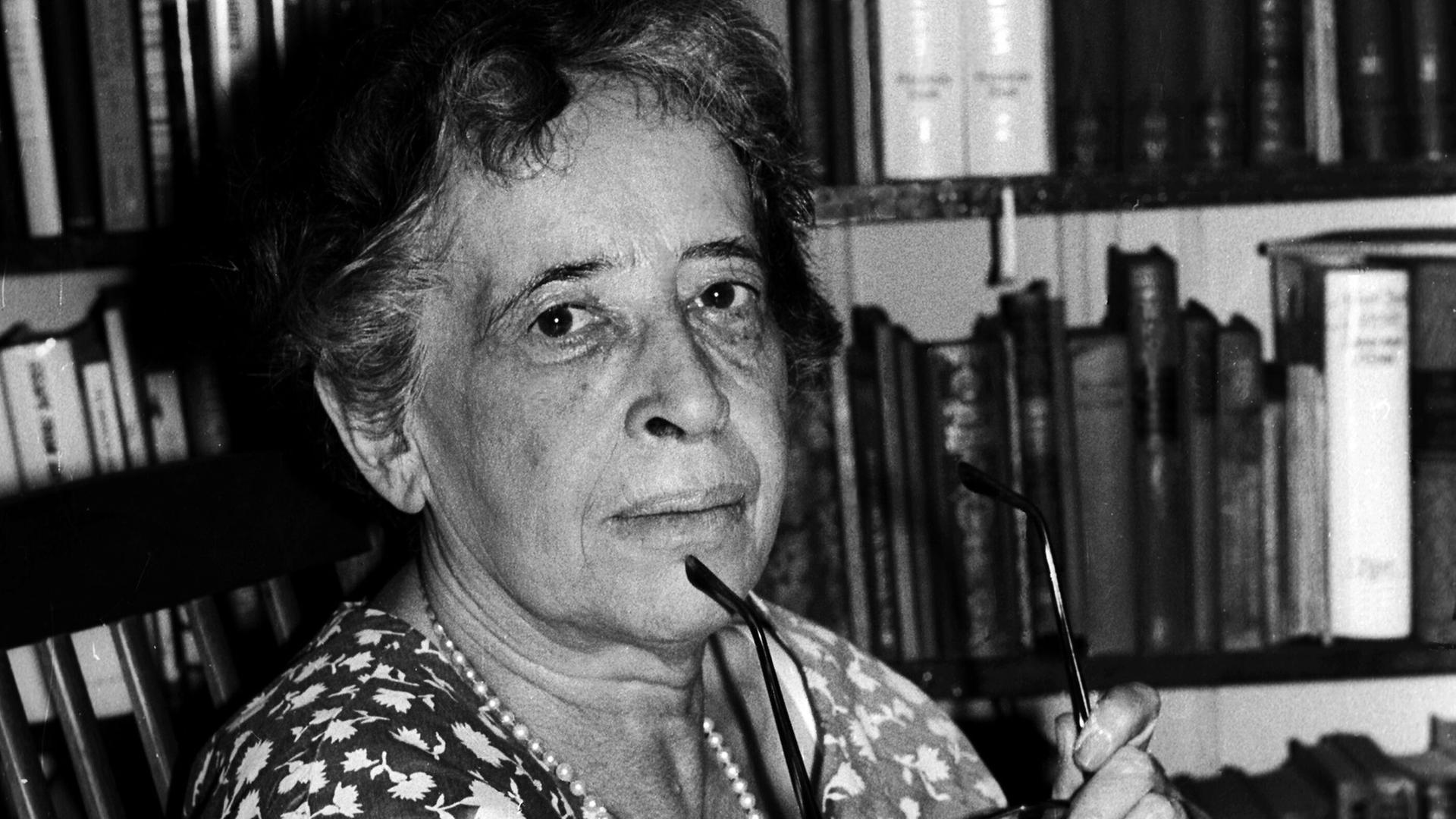 Hannah Arendt blickt auf einer Schwarz-Weiß-Fotografie, in einem Sommerkleid vor einem Bücherregal sitzend und mit einer Brille in der Hand, im Jahr 1966 in die Kamera. 