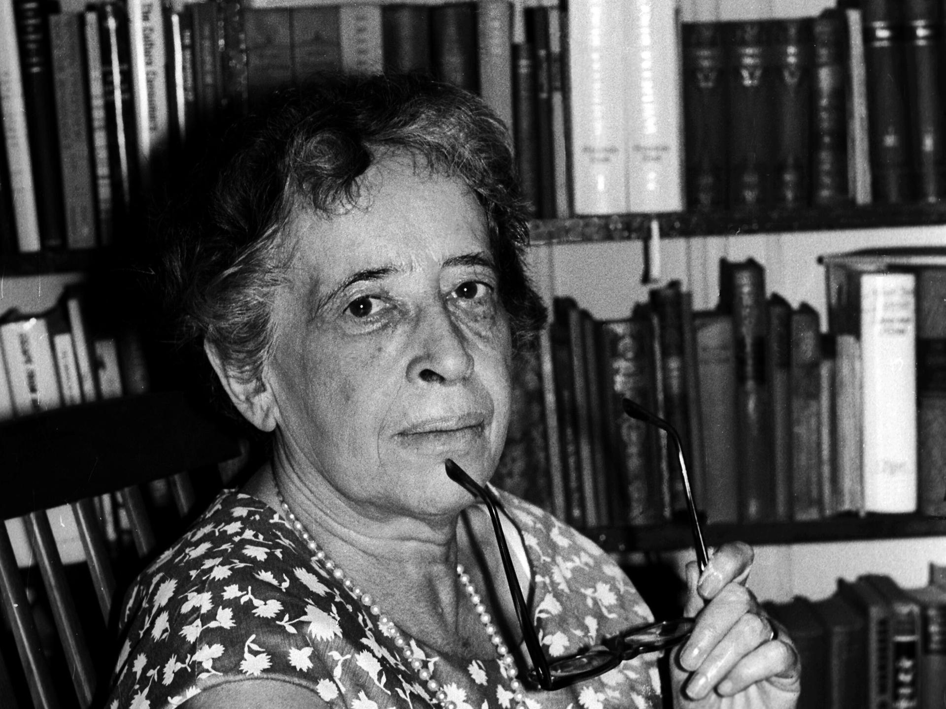 Hannah Arendt blickt auf einer Schwarz-Weiß-Fotografie, in einem Sommerkleid vor einem Bücherregal sitzend und mit einer Brille in der Hand, im Jahr 1966 in die Kamera. 