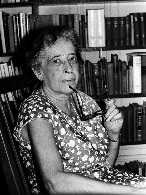 Hannah Arendt blickt auf einer Schwarz-Weiß-Fotografie, in einem Sommerkleid vor einem Bücherregal sitzend und mit einer Brille in der Hand, im Jahr 1966 in die Kamera. 