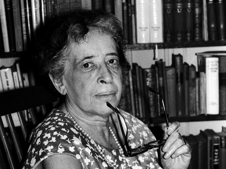 Hannah Arendt blickt auf einer Schwarz-Weiß-Fotografie, in einem Sommerkleid vor einem Bücherregal sitzend und mit einer Brille in der Hand, im Jahr 1966 in die Kamera. 