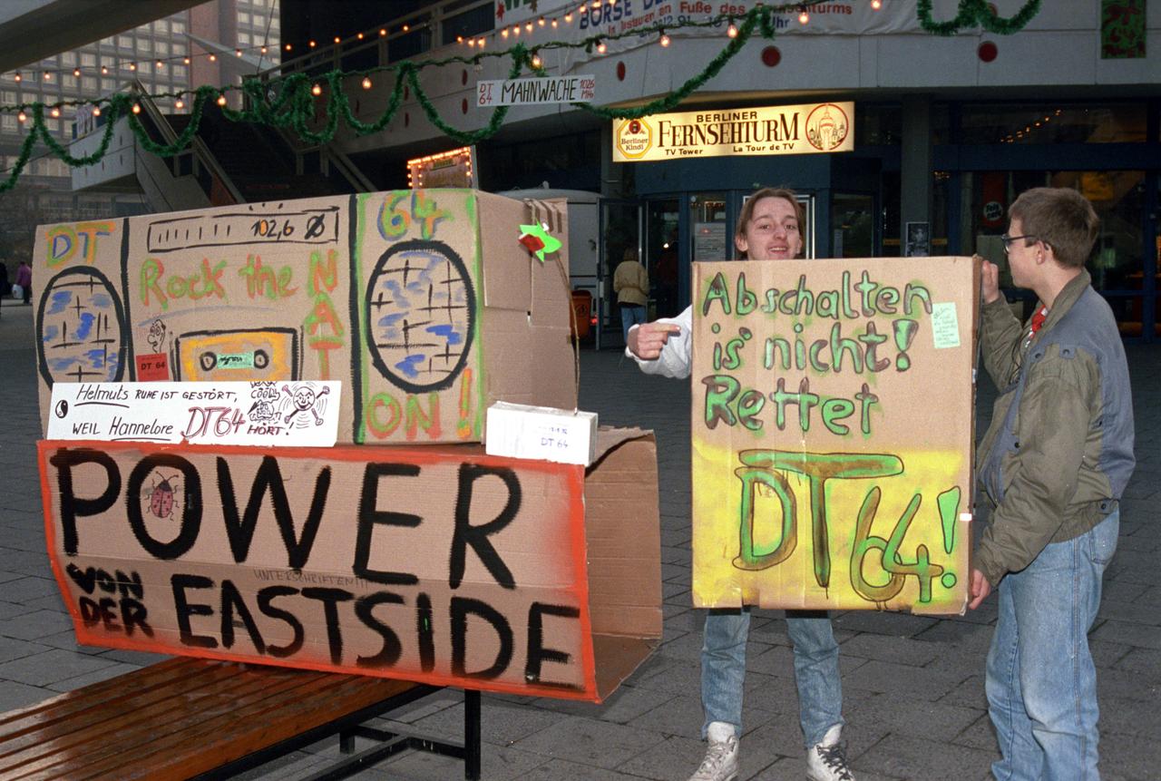 Zwei Jugendliche halten eine Mahnwache für DT 64 auf dem Berliner Alexanderplatz. Sie haben einen großen Pappkarton als Radio gestaltet. Auf einem Karton steht "Power von der Eastside". Sie halten eine Pappe mit der Aufschrift "Abschalten is nich. Rettet DT64".