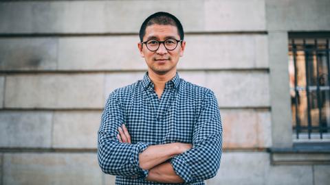 Der Architekt Van Bo Le-Mentzel steht mit verschränkten Armen vor einer Häuserwand. Er trägt ein blau-kariertes Hemd und eine schwarze, runde Brille.