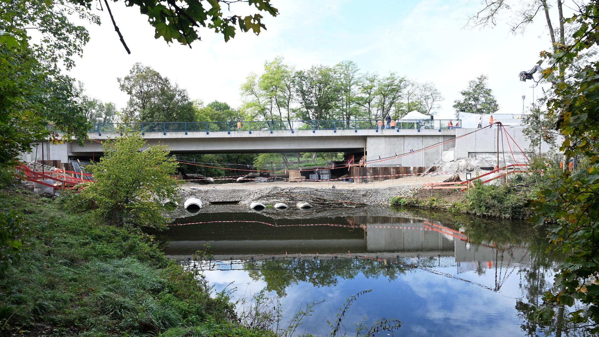 Hinter einer Baustelle ist die Struktur einer Brücke zu sehen, davor ein Fluss.