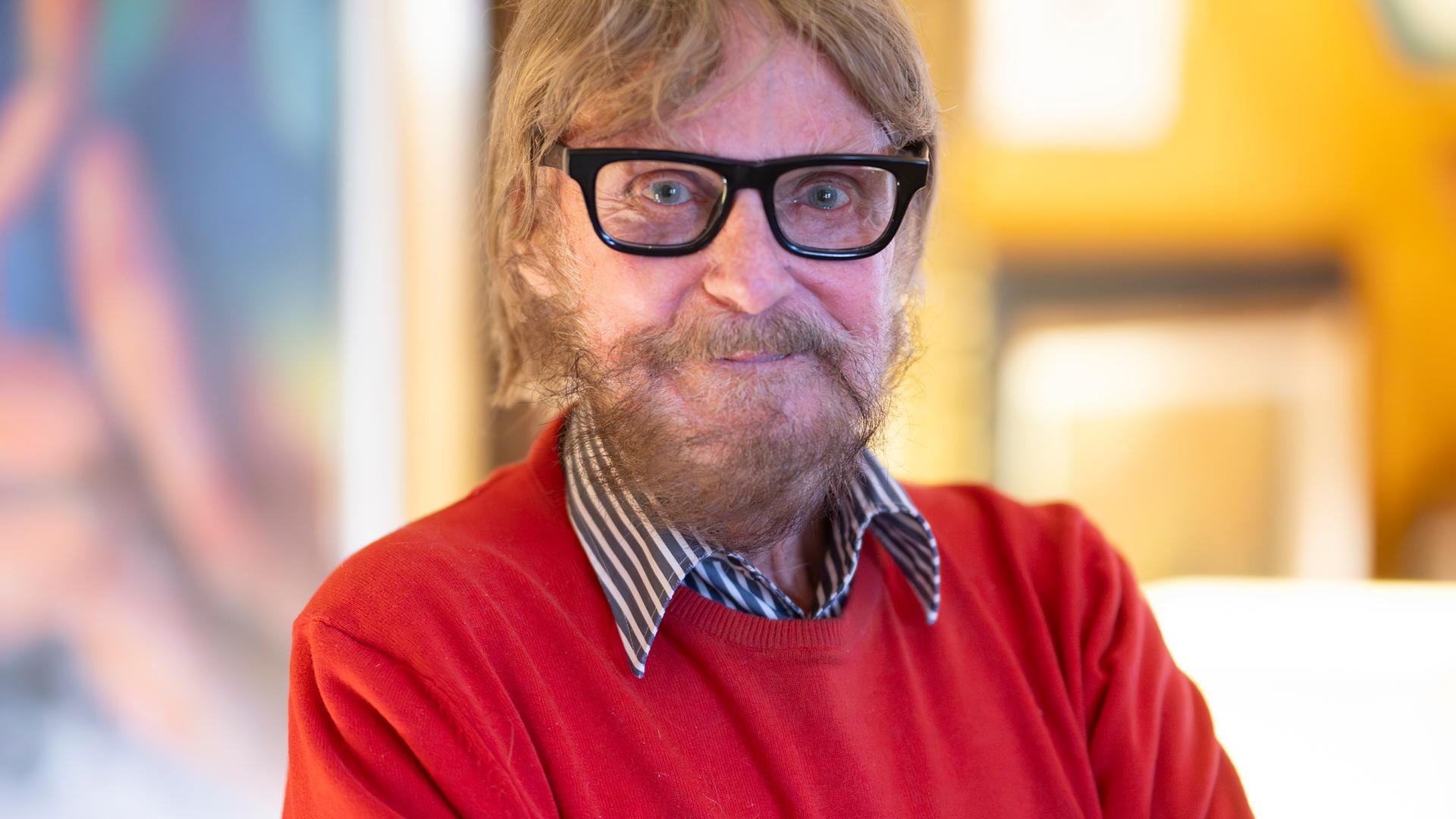 Der Kunstsammler Frank Brabant steht in seiner Wohnung in Wiesbaden. Er trägt eine schwarze Brille, einen roten Pullover und schaut freundlich in die Kamera.