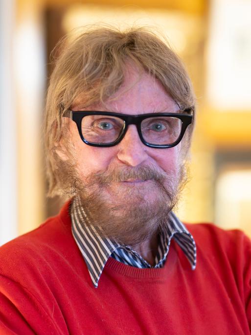 Der Kunstsammler Frank Brabant steht in seiner Wohnung in Wiesbaden. Er trägt eine schwarze Brille, einen roten Pullover und schaut freundlich in die Kamera.