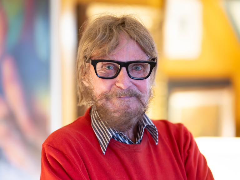 Der Kunstsammler Frank Brabant steht in seiner Wohnung in Wiesbaden. Er trägt eine schwarze Brille, einen roten Pullover und schaut freundlich in die Kamera.