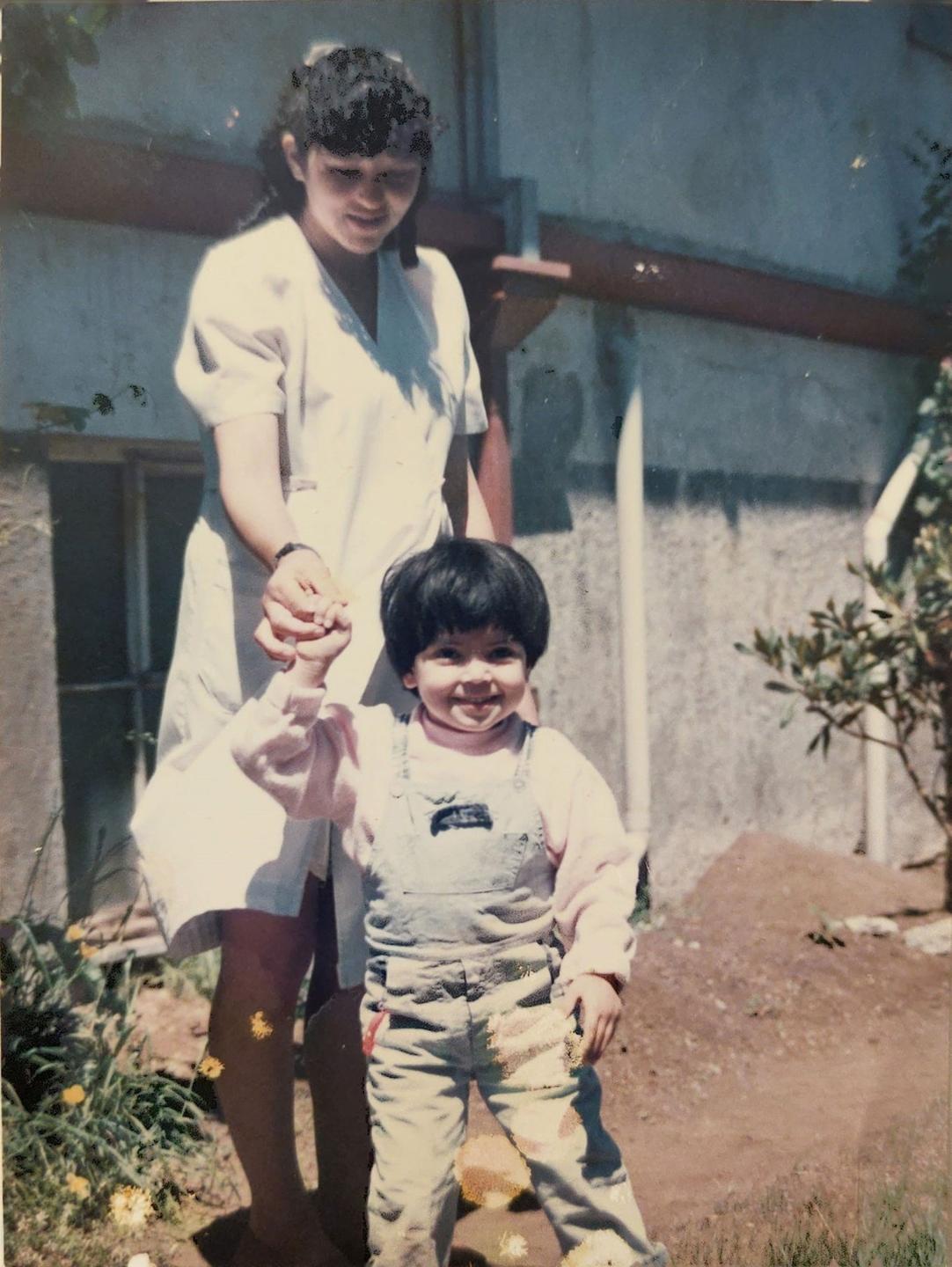 Ein altes Farbfoto zeigt ein kleines lachendes Mädchen an der Hand einer jungen Frau im weißen Kittel.