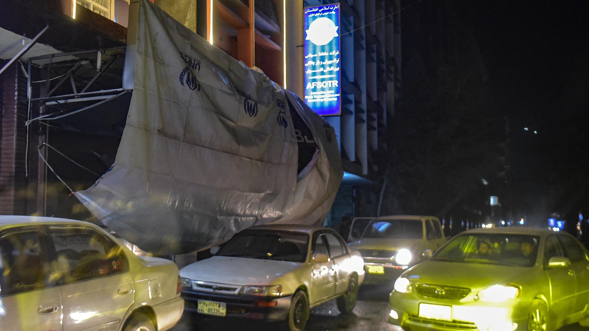 Eine Plane hängt vor dem Eingang eines Gebäudes, davor fahren Autos auf einer Straße entlang.