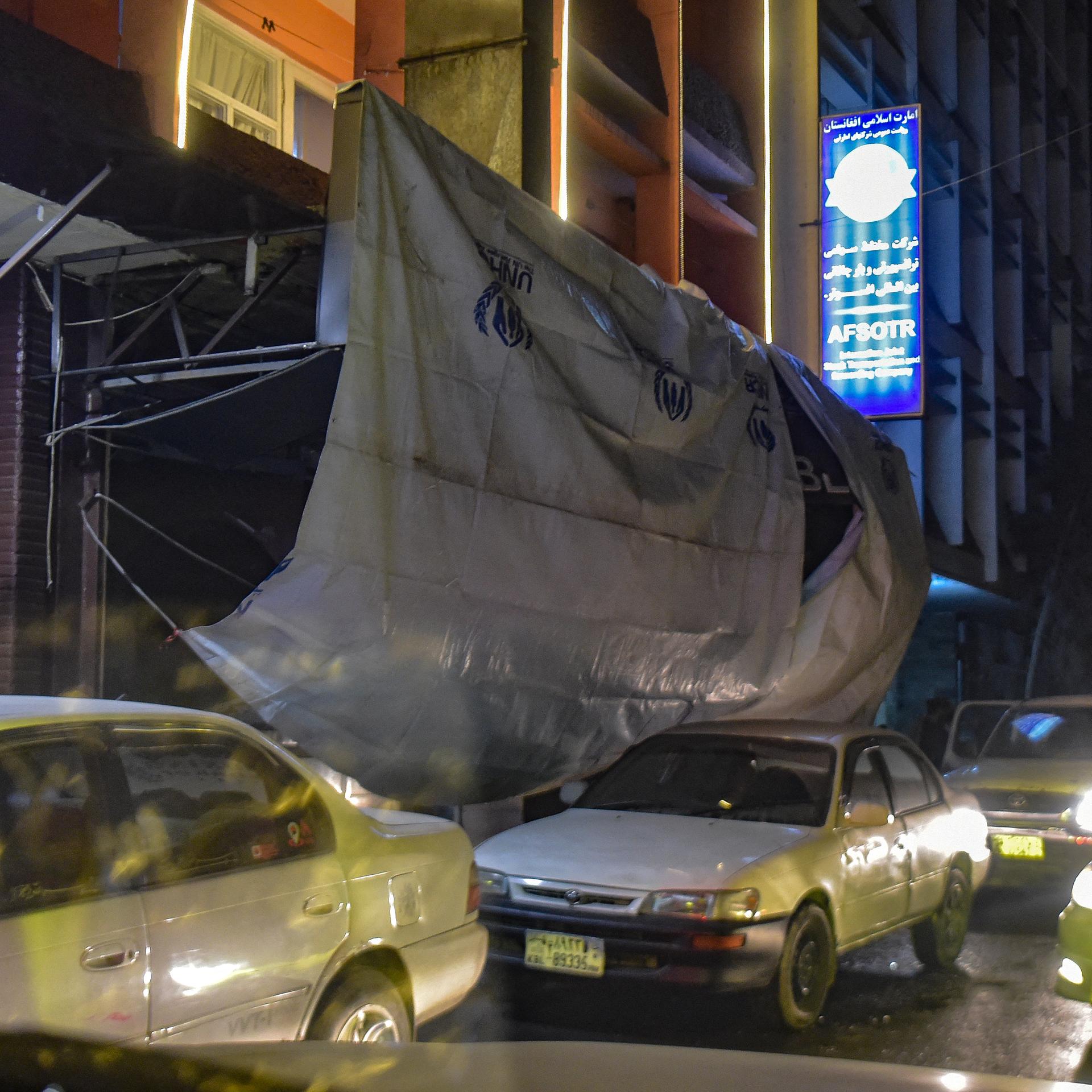 Eine Plane hängt vor dem Eingang eines Gebäudes, davor fahren Autos auf einer Straße entlang.