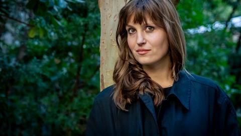 Eine Frau lehnt an einem Baum und schaut in die Kamera: die Schriftstellerin Charlotte Gneuß.