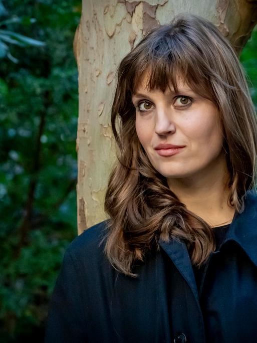 Eine Frau lehnt an einem Baum und schaut in die Kamera: die Schriftstellerin Charlotte Gneuß.