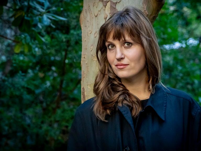 Eine Frau lehnt an einem Baum und schaut in die Kamera: die Schriftstellerin Charlotte Gneuß.