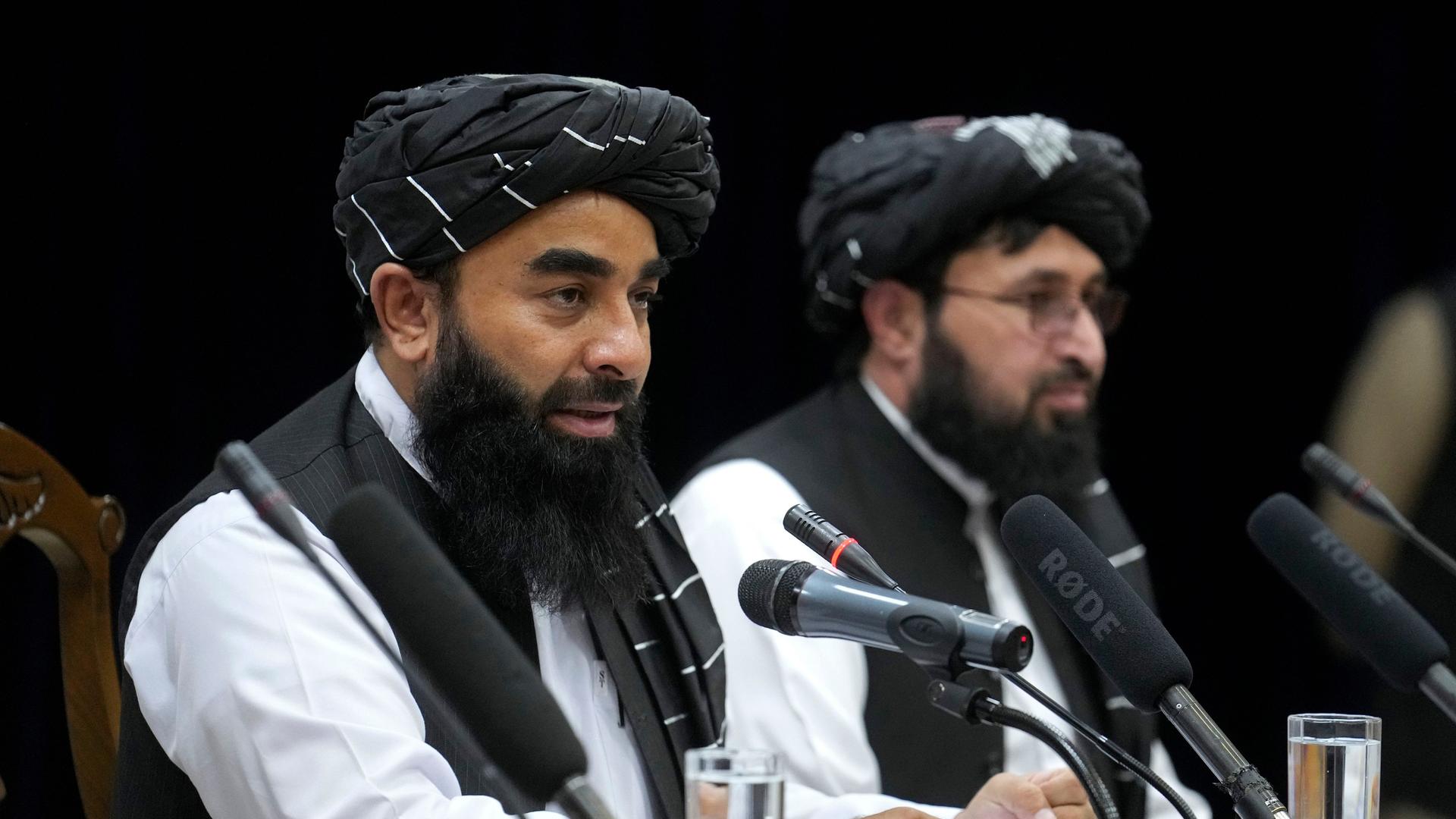 Das Foto zeigt zwei Sprecher der Taliban. Sie sitzen an einem Tisch mit Mikrofonen.