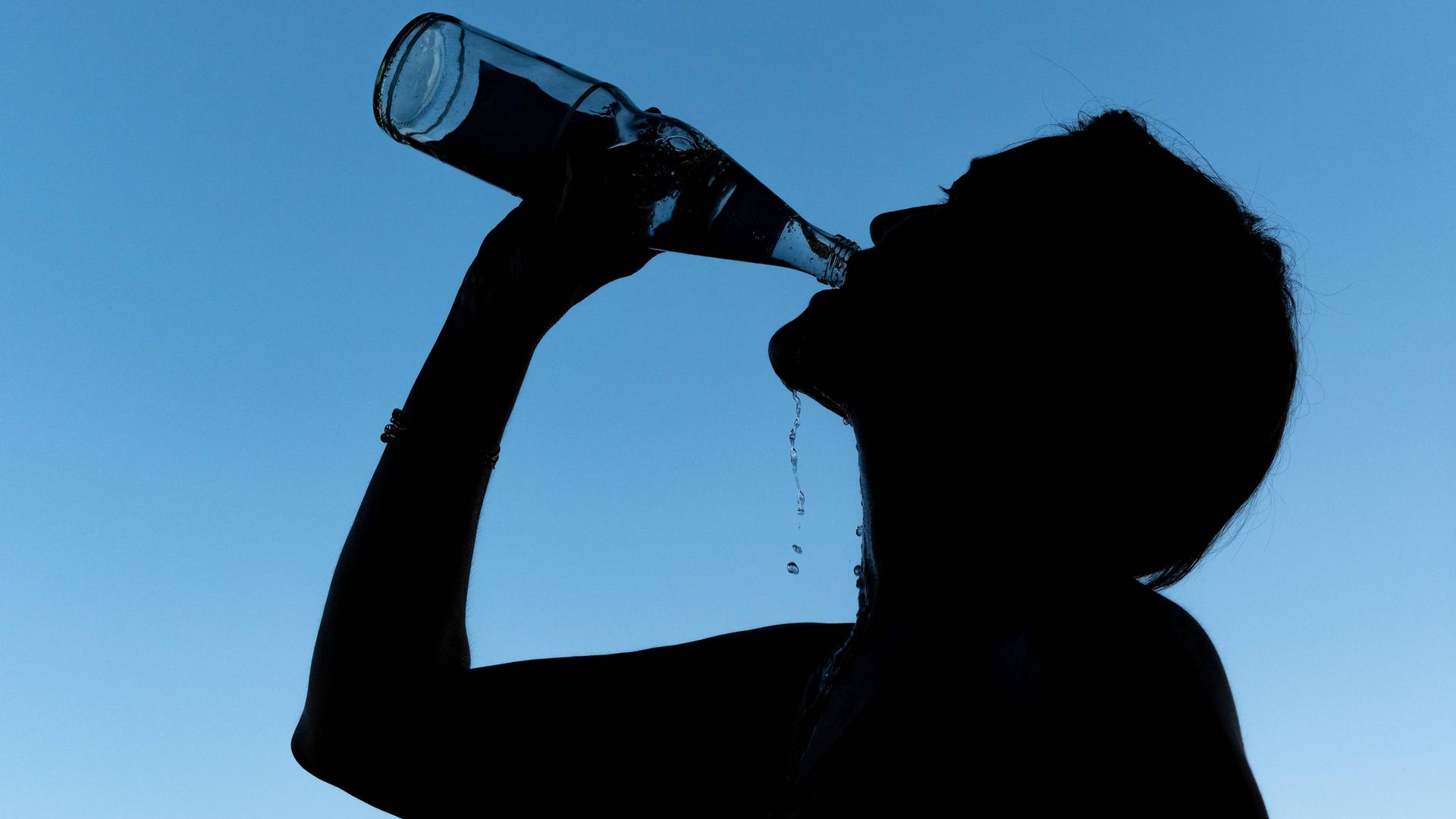 Die Silhouette einer Frau, die aus einer Mineralwasserflasche trinkt, ist vor blauem Hintergrund zu sehen. Die Silhouette einer Frau, die aus einer Mineralwasserflasche trinkt, ist vor blauem Hintergrund zu sehen.