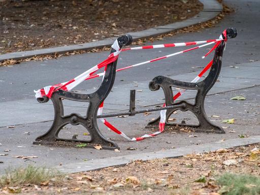 Grundgestell einer Parkbank mit Absperrband zu sehen. Der Bank fehlen Sitzfläche und Lehne.