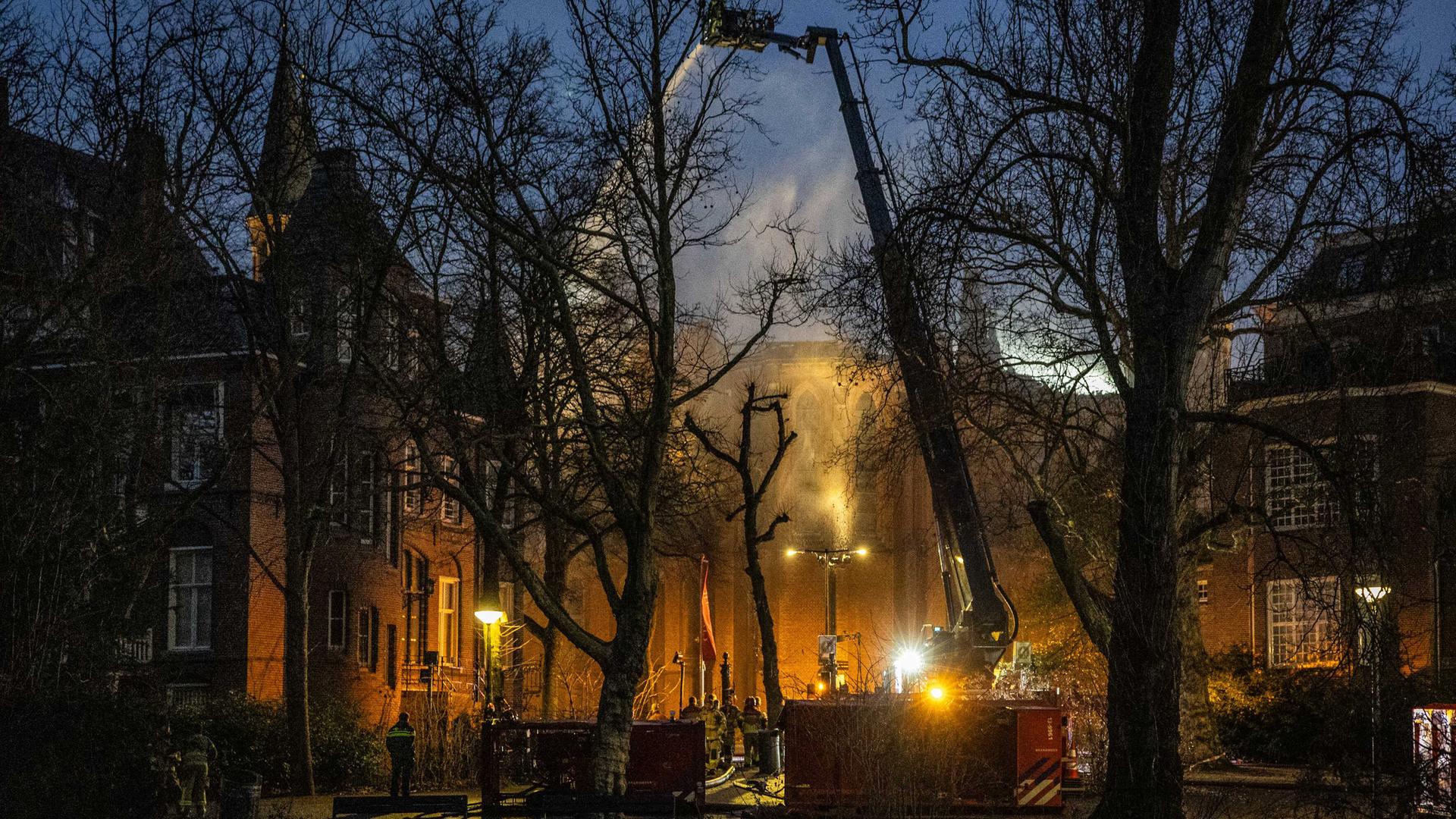 Zu sehen ist die Vondelkirche in Amsterdam, aus dem Dach steigt Rauch auf, im Vordergrund ein großer Kran der Feuerwehr.