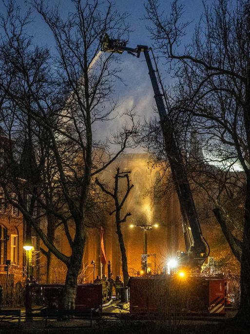 Zu sehen ist die Vondelkirche in Amsterdam, aus dem Dach steigt Rauch auf, im Vordergrund ein großer Kran der Feuerwehr.