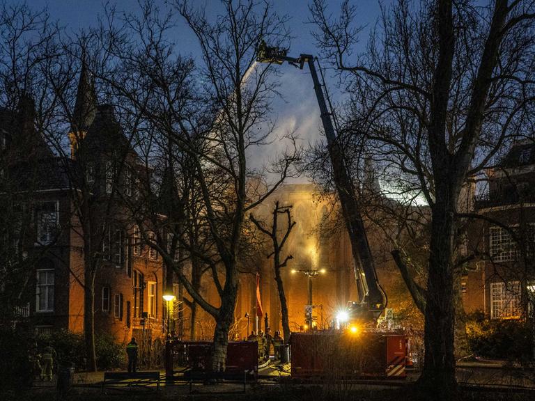 Zu sehen ist die Vondelkirche in Amsterdam, aus dem Dach steigt Rauch auf, im Vordergrund ein großer Kran der Feuerwehr.