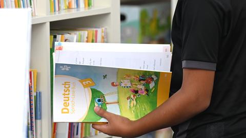 Ein Schulkind steht vor einem Regal und hält ein Arbeitsheft "Deutsch" in der Hand. Symbolbild