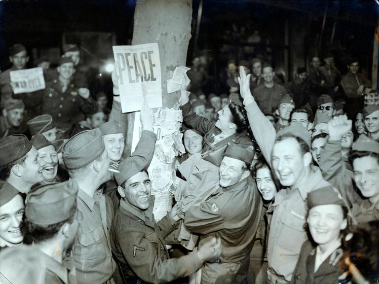 Lachende, feiernde amerikanische Soldaten. Einer hält eine Zeitung hoch, auf der in Großbuchstaben "Peace" gedruckt ist.