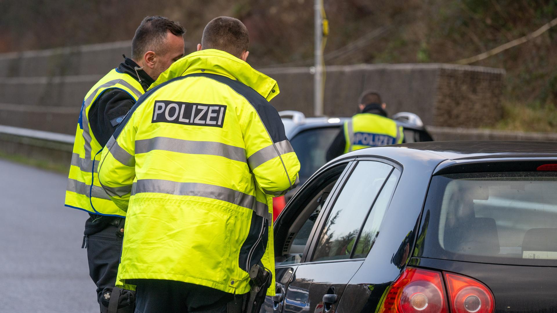 Polizisten stehen auf einer Straße. Vor den Polizisten steht ein Auto. Eine Fenster-Scheibe von dem Auto ist offen. Polizisten stehen auf einer Straße. Vor den Polizisten steht ein Auto. Eine Fenster-Scheibe von dem Auto ist offen.
