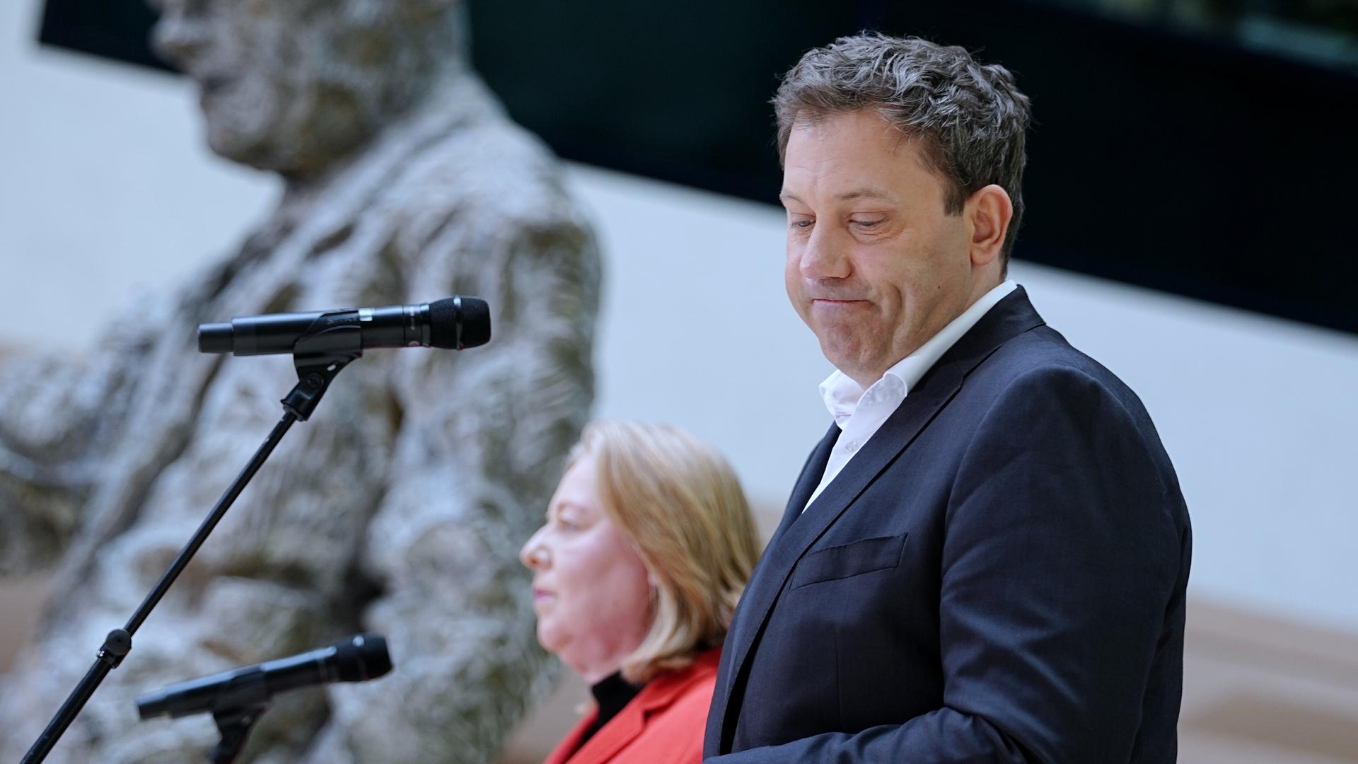 Ein Mann im Anzug steht im Vordergrund an einem Mikrofon und blickt mit ernstem Gesicht zur Seite. Hinter ihm steht eine Frau in einem roten Anzug an einem zweiten Mikrofon. Beide sind auf einer Bühne zu sehen. 