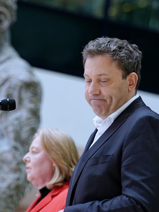 Ein Mann im Anzug steht im Vordergrund an einem Mikrofon und blickt mit ernstem Gesicht zur Seite. Hinter ihm steht eine Frau in einem roten Anzug an einem zweiten Mikrofon. Beide sind auf einer Bühne zu sehen. 