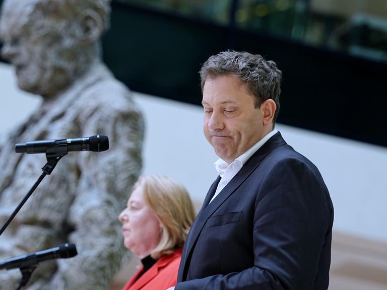 Ein Mann im Anzug steht im Vordergrund an einem Mikrofon und blickt mit ernstem Gesicht zur Seite. Hinter ihm steht eine Frau in einem roten Anzug an einem zweiten Mikrofon. Beide sind auf einer Bühne zu sehen. 