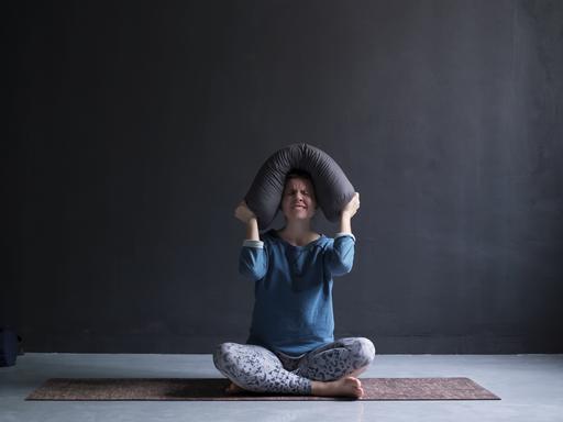 Eine Frau sitzt auf einer Yogamatte und drückt wütend ein Kissen auf ihren Kopf.