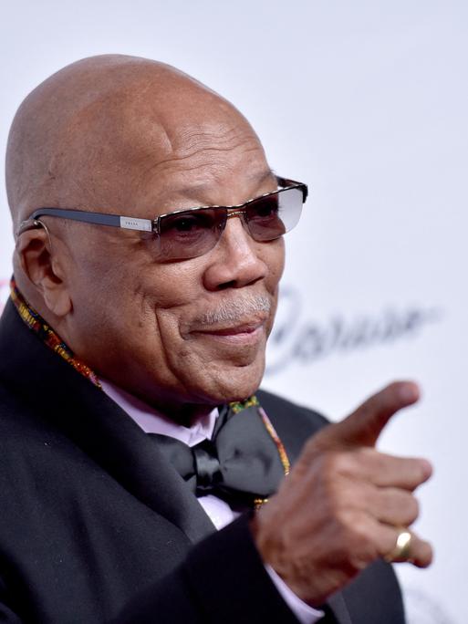 Quincy Jones mit Hemd, Anzug, Fliege und Sonnenbrille vor hellem Hintergrund. Er hebt den Zeigefinger der rechten Hand in die Luft.