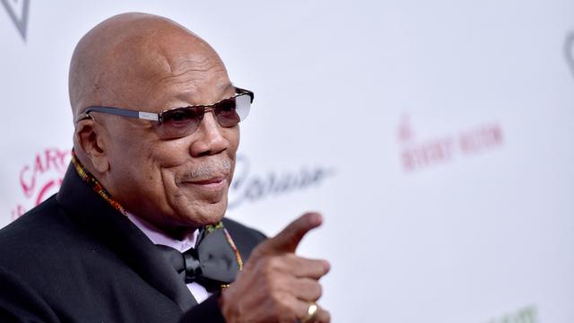 Quincy Jones mit Hemd, Anzug, Fliege und Sonnenbrille vor hellem Hintergrund. Er hebt den Zeigefinger der rechten Hand in die Luft.