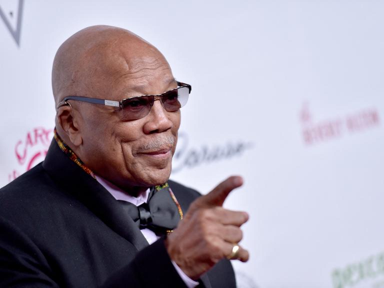 Quincy Jones mit Hemd, Anzug, Fliege und Sonnenbrille vor hellem Hintergrund. Er hebt den Zeigefinger der rechten Hand in die Luft.