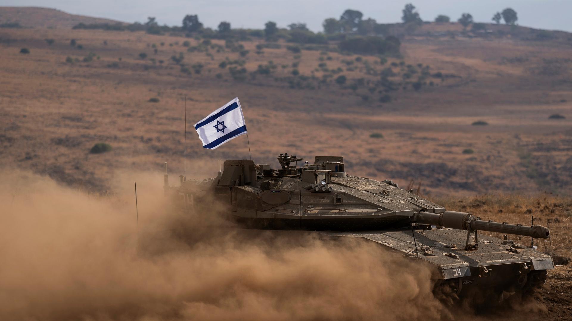Der Panzer mit einer israelischen Flagge auf dem Dach fährt durch eine braune hügelige Ebene und wirbelt Staub auf.