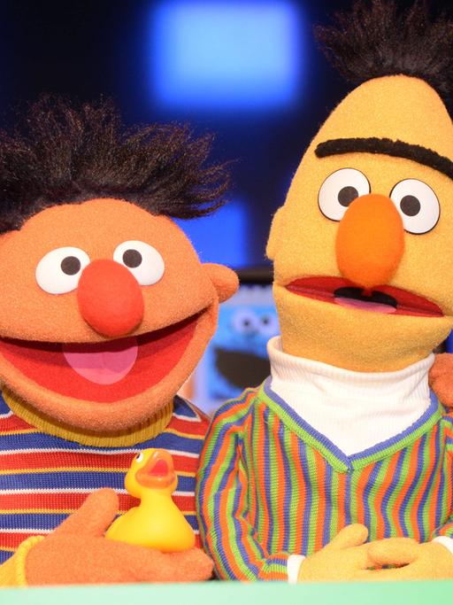 Die Puppen Ernie & Bert aus der Sesamstraße. Ernie hat ein Quietscheentchen in der Hand und seien Arm um Bert gelegt