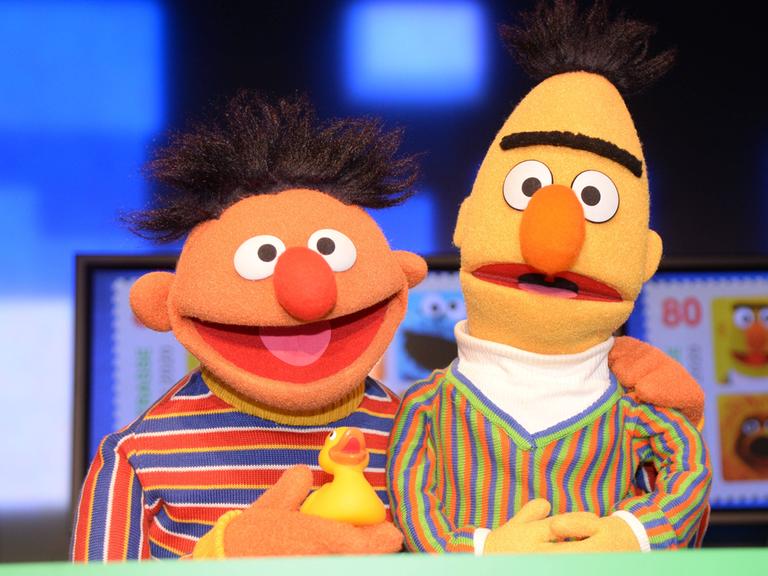 Die Puppen Ernie & Bert aus der Sesamstraße. Ernie hat ein Quietscheentchen in der Hand und seien Arm um Bert gelegt