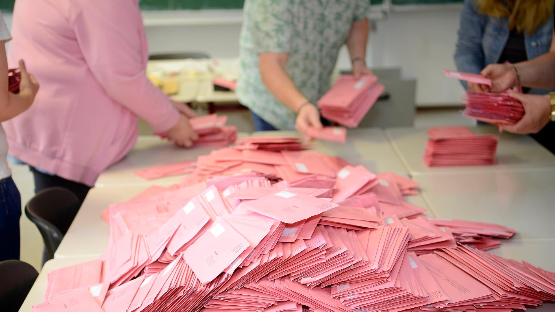 Viele rosa Wahlumschläge liegen auf einem Tisch. Menschen stehen an dem Tisch und sortieren die Umschläge.