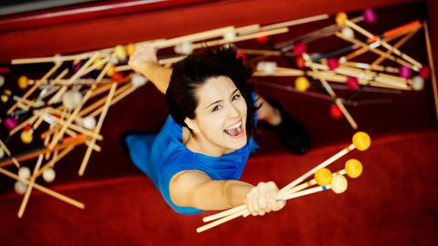 Eine Frau im blauen Kleid lacht strahlend in die Kamera. Sie ist von oben fotografiert und hält in der Hand mehrere Marimbaschlägel. Im Hintergrund sieht man noch einen ganzen Haufen solcher Schlägel, die wie Mikados auf dem Boden liegen.