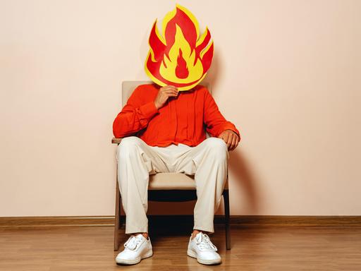 Eine Person sitzt auf einem Stuhl und hält sich eine große, aus Papier ausgeschnittene Flamme in Rot-Gelb vors Gesicht. Eine Person sitzt auf einem Stuhl und hält sich eine große, aus Papier ausgeschnittene Flamme in Rot-Gelb vors Gesicht.