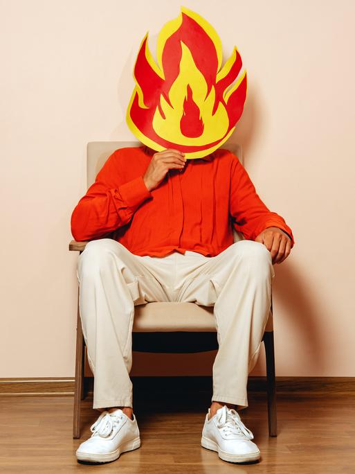 Eine Person sitzt auf einem Stuhl und hält sich eine große, aus Papier ausgeschnittene Flamme in Rot-Gelb vors Gesicht.