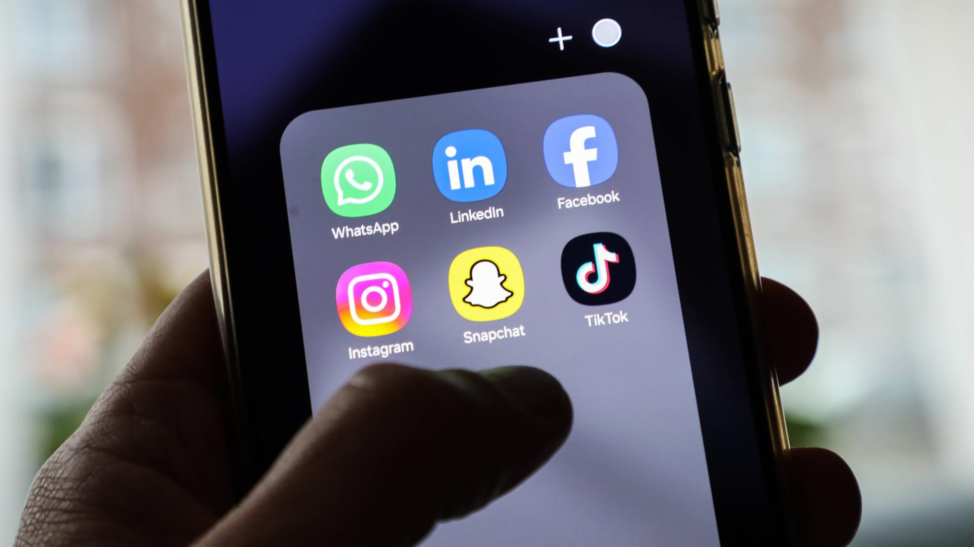 Auf einem Handybildschirm sind verschiedene Apps für Social-Media-Plattformen wie Instagram, Facebook oder Tiktok sichtbar. Über dem Bildschirm schwebt eine Hand.  Auf einem Handybildschirm sind verschiedene Apps für Social-Media-Plattformen wie Instagram, Facebook oder Tiktok sichtbar. Über dem Bildschirm schwebt eine Hand.