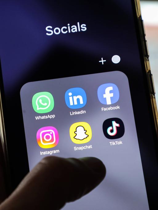 Auf einem Handybildschirm sind verschiedene Apps für Social-Media-Plattformen wie Instagram, Facebook oder Tiktok sichtbar. Über dem Bildschirm schwebt eine Hand. 