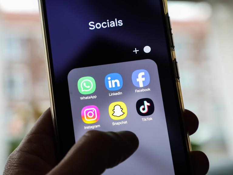 Auf einem Handybildschirm sind verschiedene Apps für Social-Media-Plattformen wie Instagram, Facebook oder Tiktok sichtbar. Über dem Bildschirm schwebt eine Hand. 