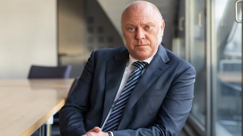 Ulrich Schneider stützt sich auf der Lehne eines Stuhls ab und schaut nachdenklich in die Kamera. Er trägt einen dunkelblauen Anzug und blau gestreifte Krawatte. Ulrich Schneider stützt sich auf der Lehne eines Stuhls ab und schaut nachdenklich in die Kamera. Er trägt einen dunkelblauen Anzug und blau gestreifte Krawatte.