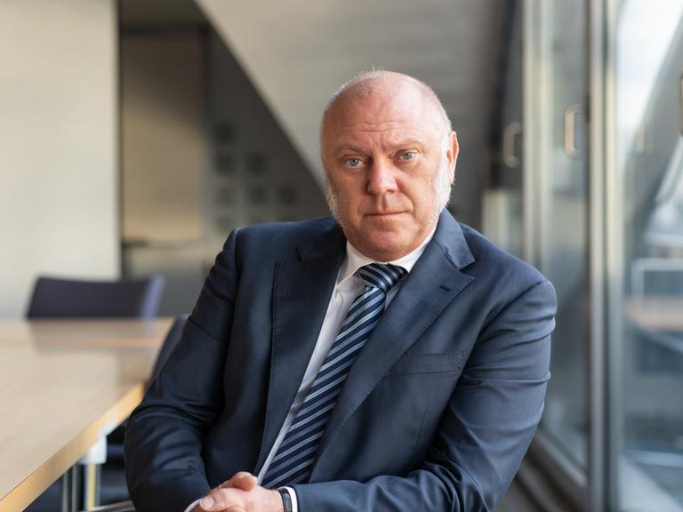 Ulrich Schneider stützt sich auf der Lehne eines Stuhls ab und schaut nachdenklich in die Kamera. Er trägt einen dunkelblauen Anzug und blau gestreifte Krawatte.