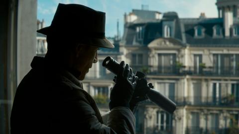 Eine Szene aus dem Netlix-Film "The Killer" in dem Michael Fassbender mit Hut und Mantel am Fenster steht und mit einem Fernglas auf die Straße runterschaut.